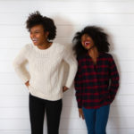 Two dark-haired young women stand against a white wall smiling and laughing while wearing long-sleeved sweaters