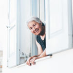 Elderly woman in need of oral care looks out the window of a white building with white shutters