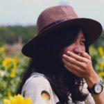 Brunette woman wearing a brown hat and watch covers her yellow teeth while standing in a sunflower field