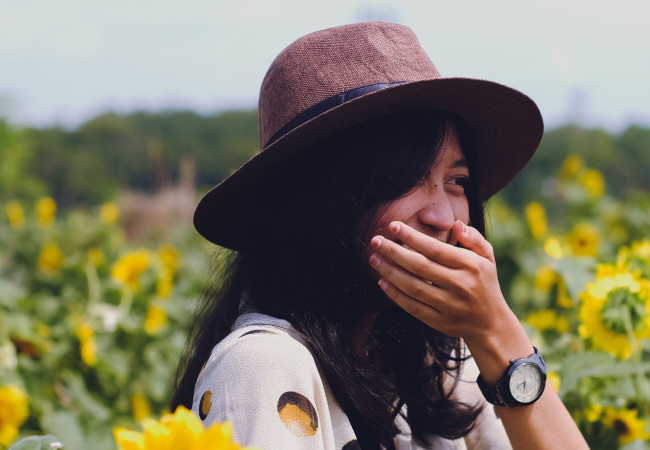 Brunette woman wearing a brown hat and watch covers her yellow teeth while standing in a sunflower field